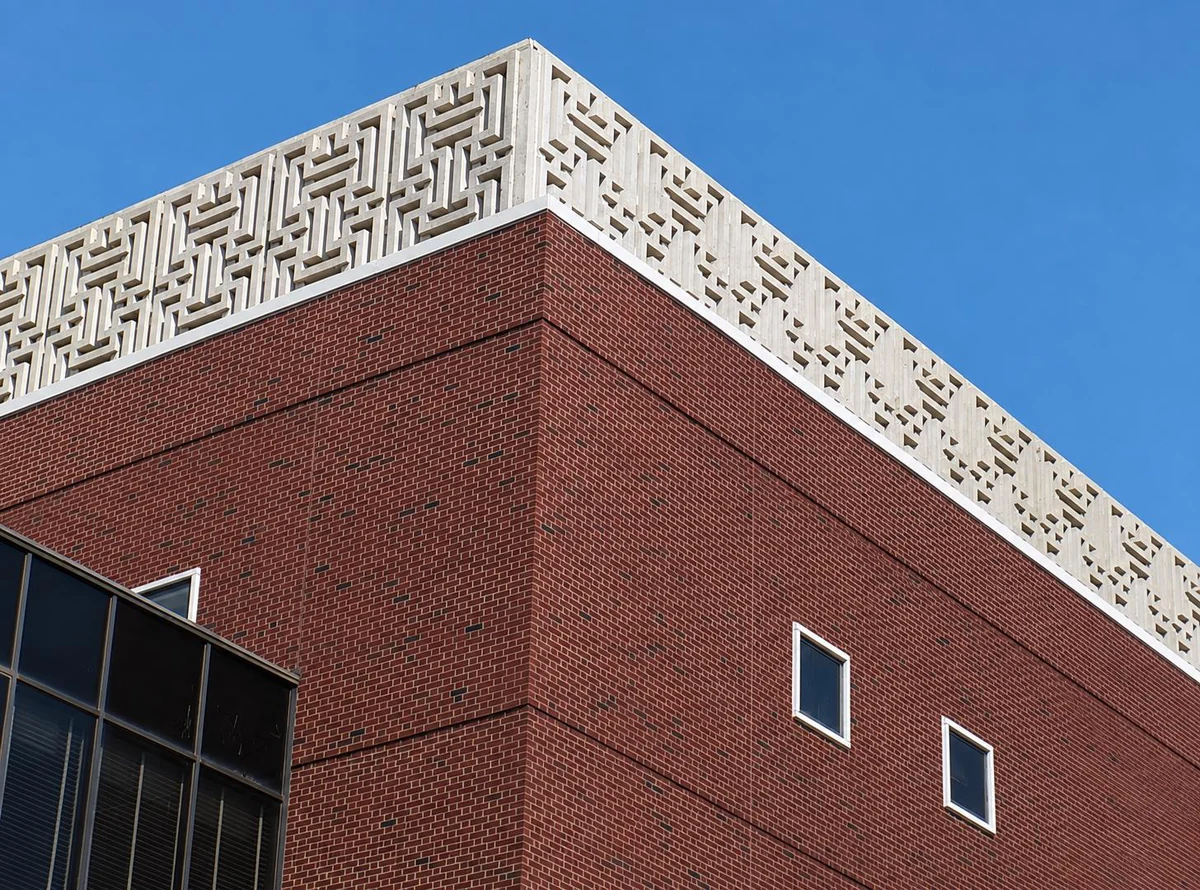 Upward view of a red brick building corner topped with a white geometric concrete lattice cornice against a clear sky