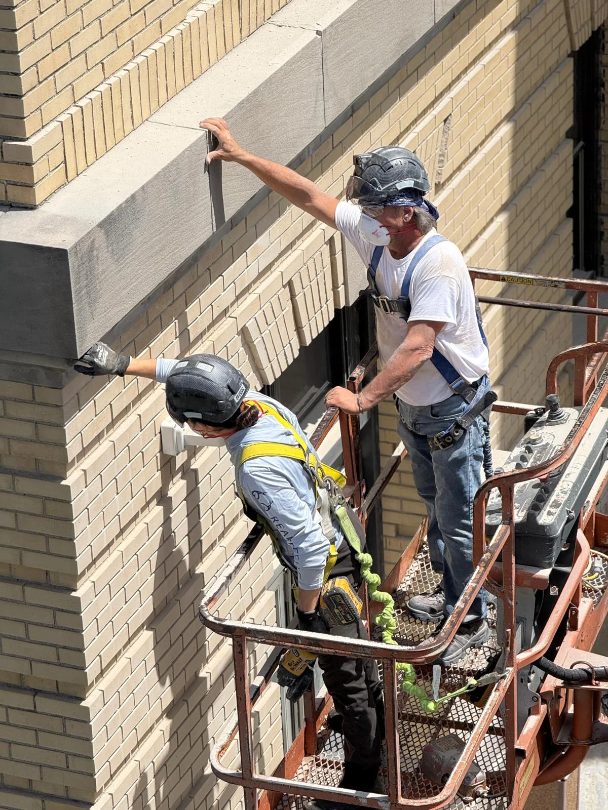 Two Bramwell-McKay technicians in a boom lift basket, harnessed and helmeted, working along the cast stone band of a cream brick building