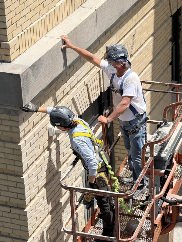 Two Bramwell-McKay technicians in a boom lift basket, harnessed and helmeted, working along the cast stone band of a cream brick building