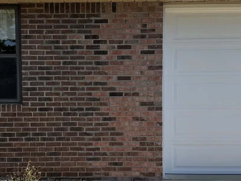 A house wall part-way through brick staining, the courses beside the garage still the original pale pink and the wall left of them already an even red brown
