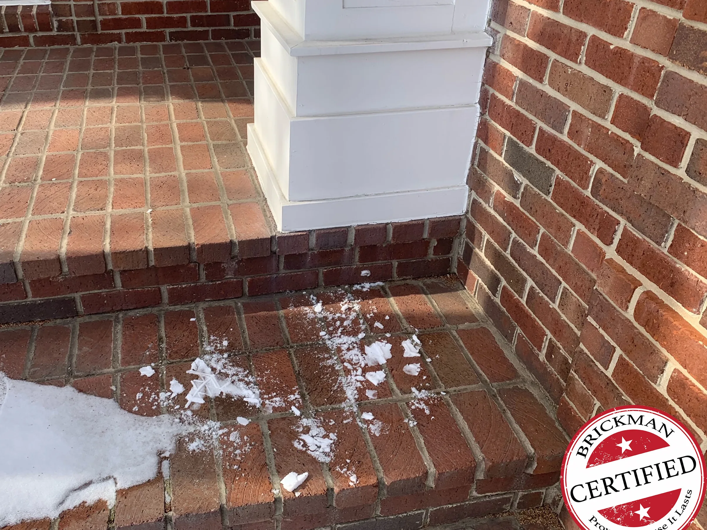 The porch pavers and top step at the base of a white column in 2021, with snow on the lower tread and the certification stamp in the corner