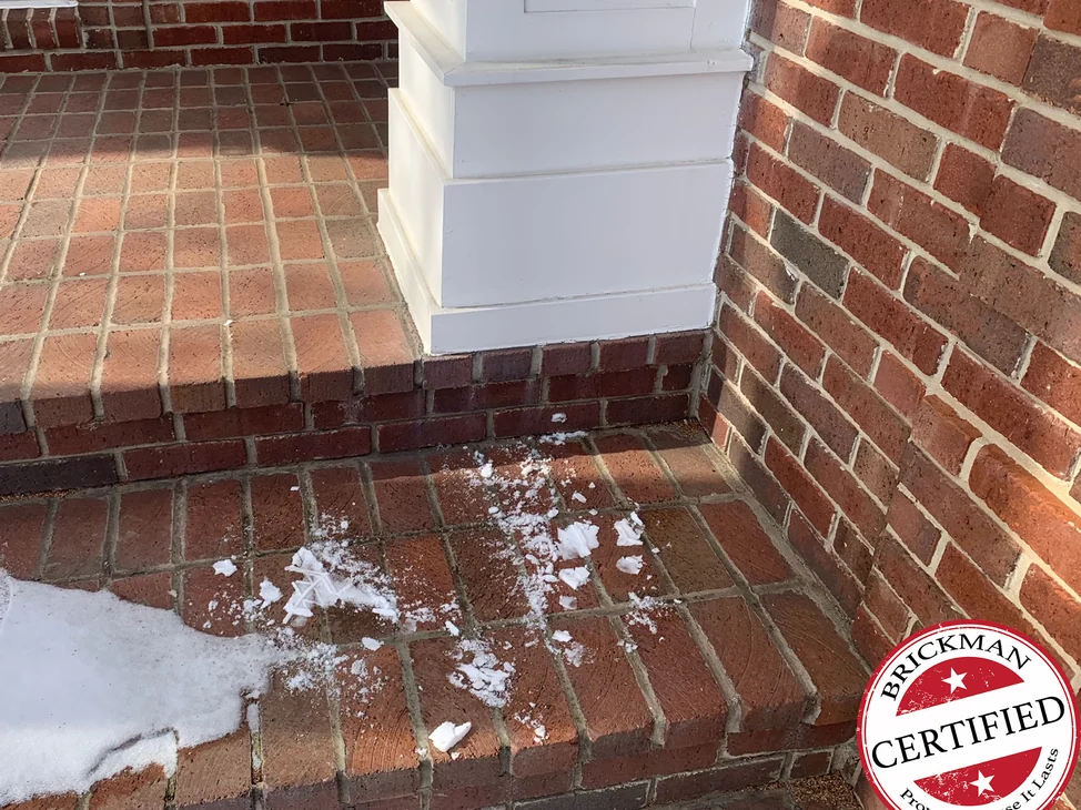 The porch pavers and top step at the base of a white column in 2021, with snow on the lower tread and the certification stamp in the corner