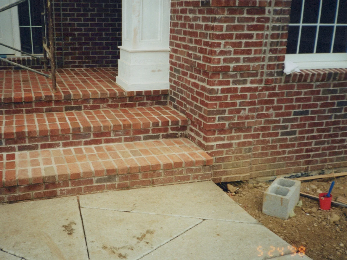 The brick steps and porch of the house entrance in May 1998 with the work underway, the lowest tread still lighter than the treads above it, a bucket and brush on the ground beside the walk
