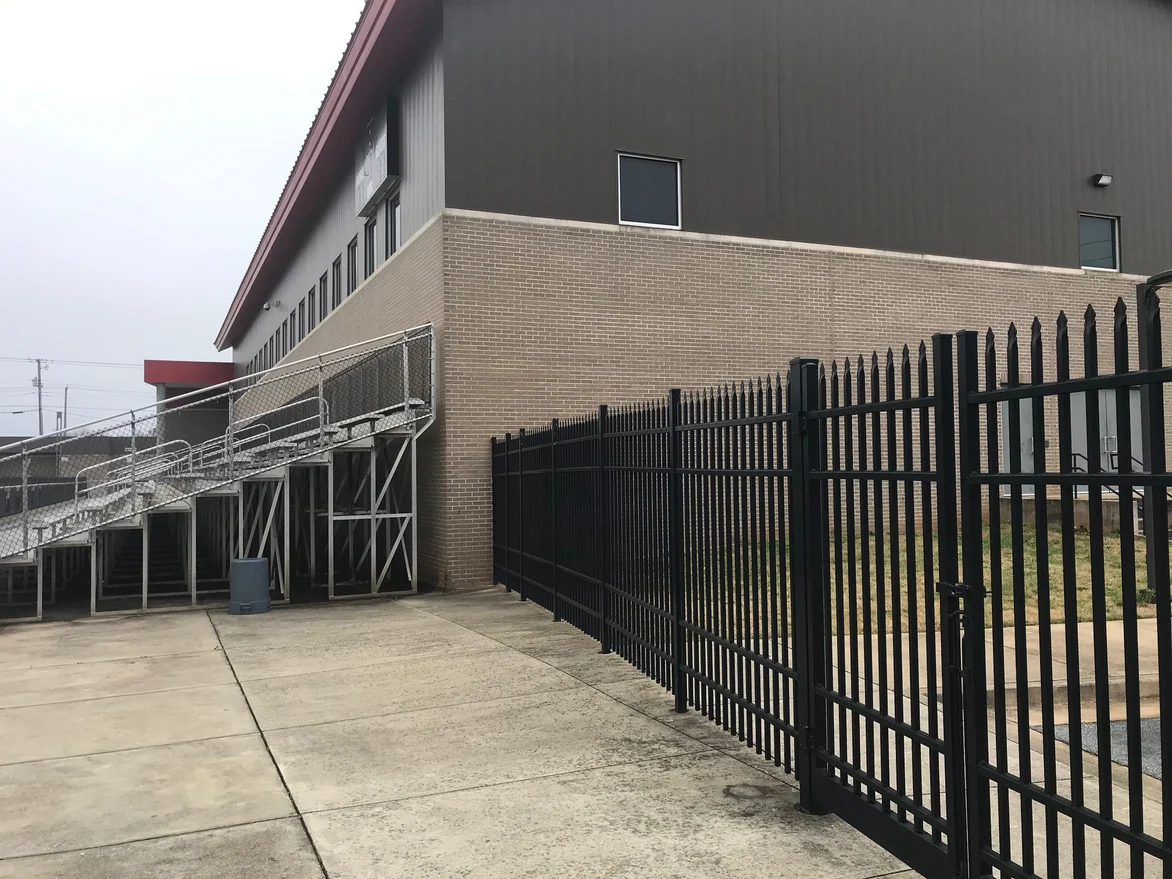 A corner of the fieldhouse in 2021 seen past a black steel fence, tan brick with pale mortar joints running back toward the bleachers