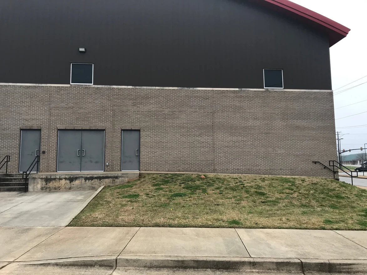 The street side of the fieldhouse in 2021, a long tan brick wall with pale mortar joints under dark metal cladding, three grey doors at the left