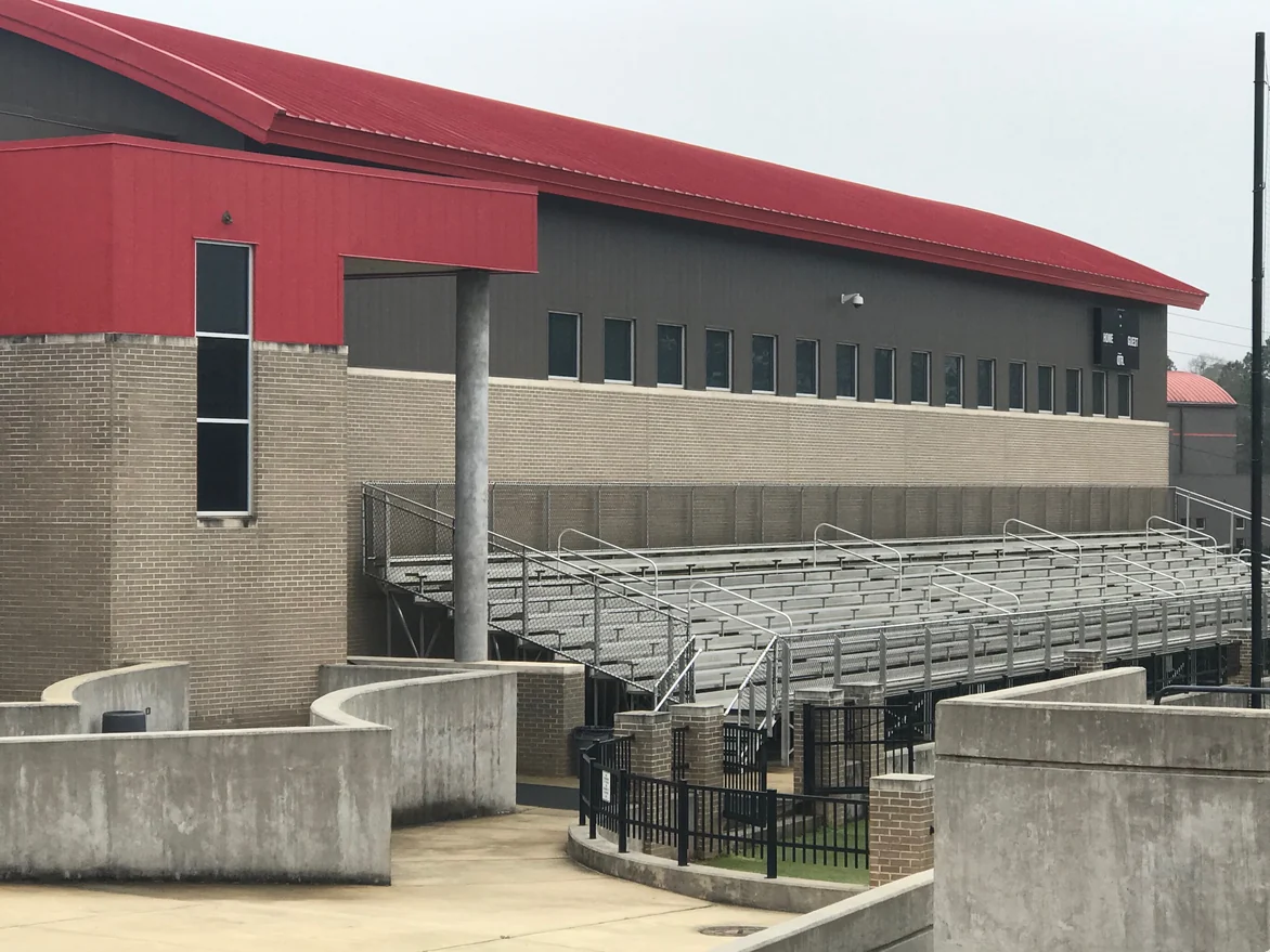 The fieldhouse and the bleachers in 2021, a band of tan brick with pale mortar joints under dark metal cladding and a red roof
