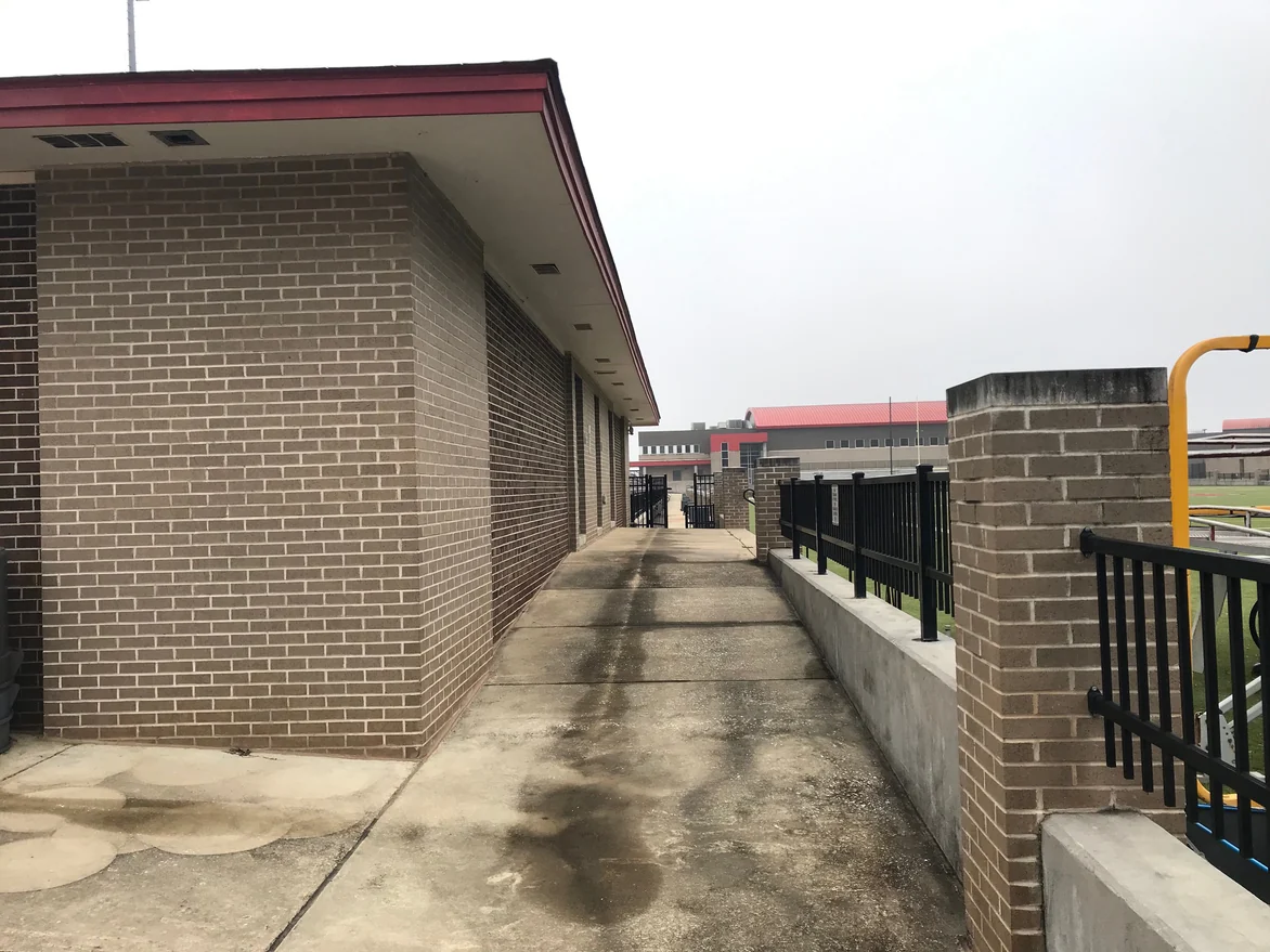 A walkway beside a stadium building in 2021, tan brick walls and fence piers with pale mortar joints, the school buildings beyond