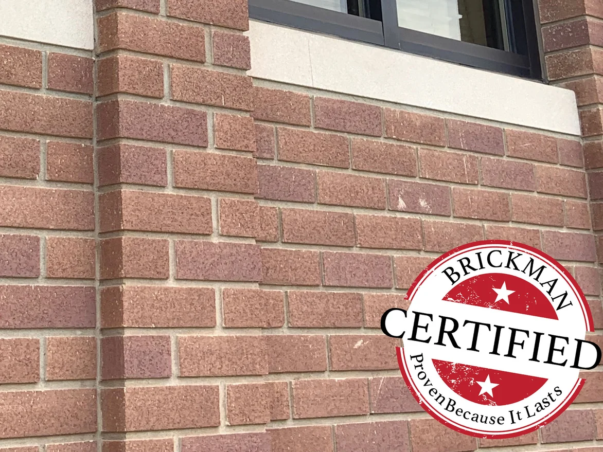 An outside corner of the wall in 2019 under a cast stone sill band, the brick even on both faces, with the Brickman certified stamp in the corner of the frame