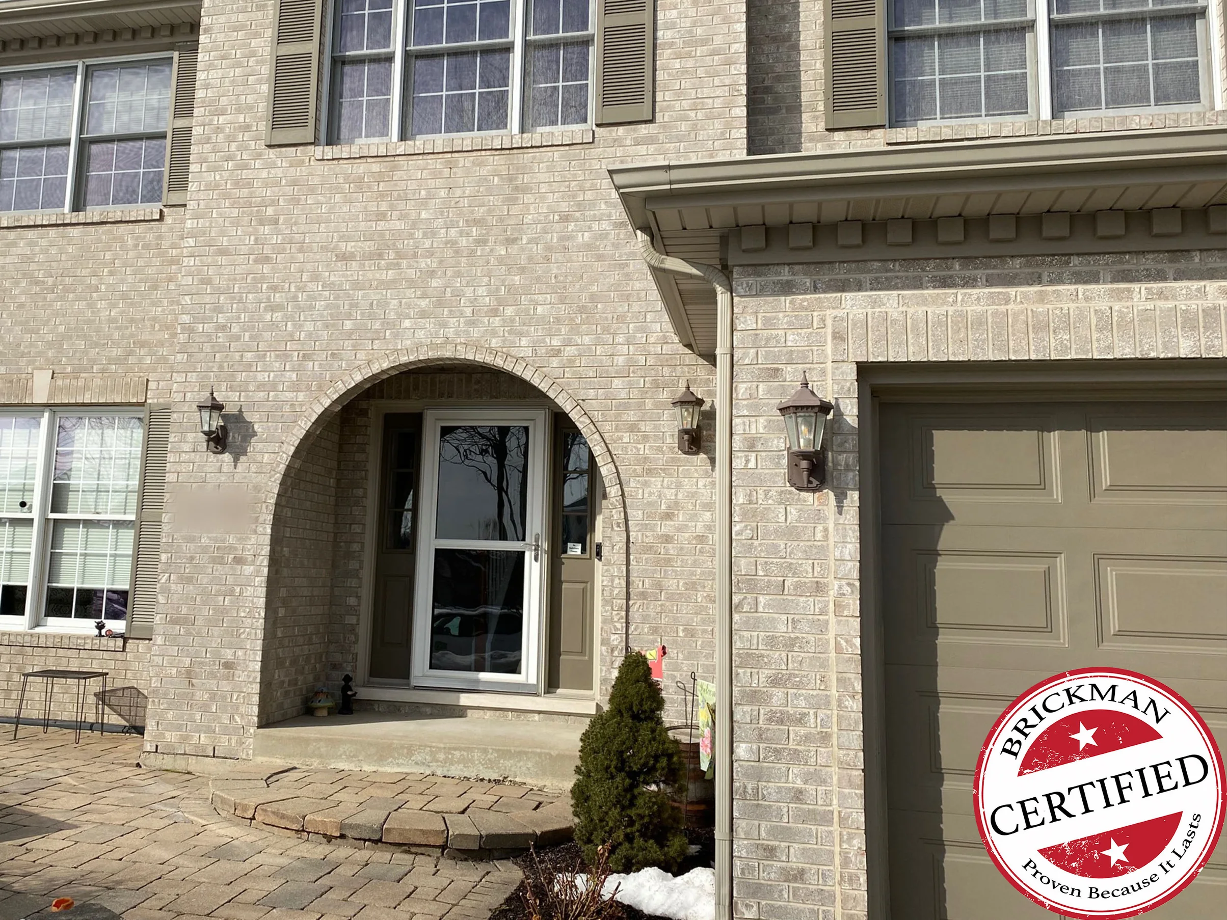 The entry of the house in 2021, tan brick around the arched front door with a lantern each side, the garage door beside it, and the certification stamp in the corner