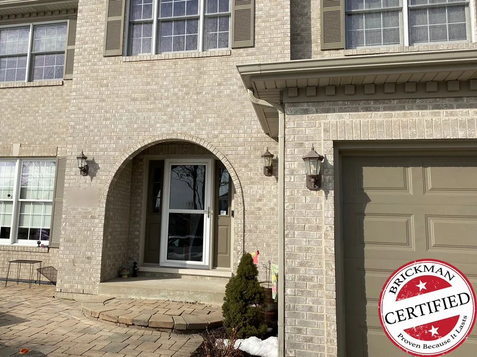 The entry of the house in 2021, tan brick around the arched front door with a lantern each side, the garage door beside it, and the certification stamp in the corner.