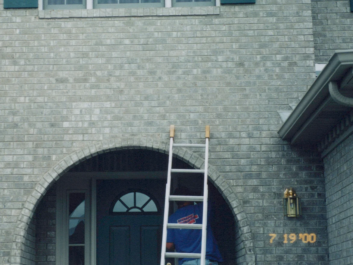 An applicator in a blue shirt on an extension ladder set inside the arched entry in July 2000, grey brick above him with the camera date burned into the corner