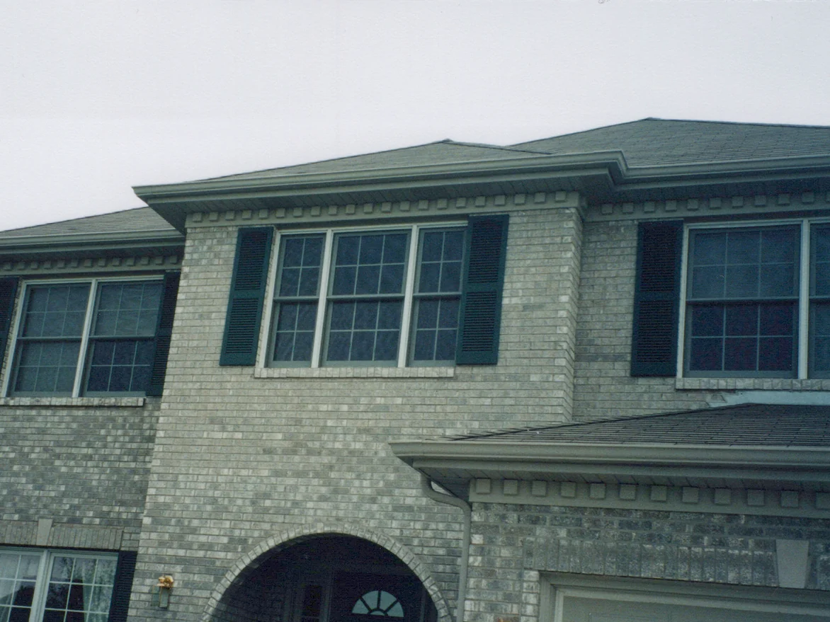 The front of the two-story house in 2000 under an overcast sky, grey brick with lighter and darker units scattered across the upper wall, dark shutters at three windows and an arched entry below
