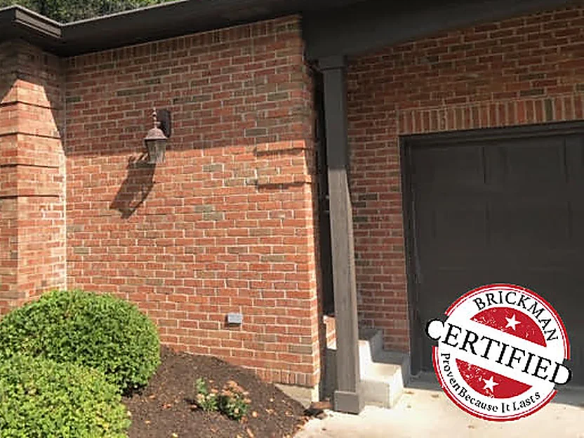An outside corner of the house in 2019 with a carriage lamp on the wall and a stepped brick pier at the angle, the brick an even red and brown range, the Brickman certified stamp in the bottom corner of the frame