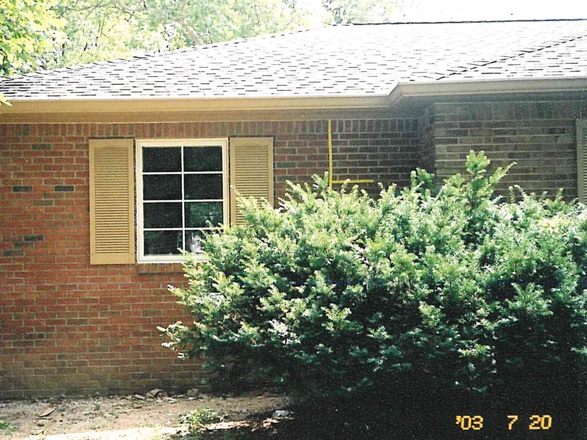The corner of a single-story brick house in 2003, red brick running under a wide hipped eave beside a shuttered window, with a yellow crayon mark on the wall to the right