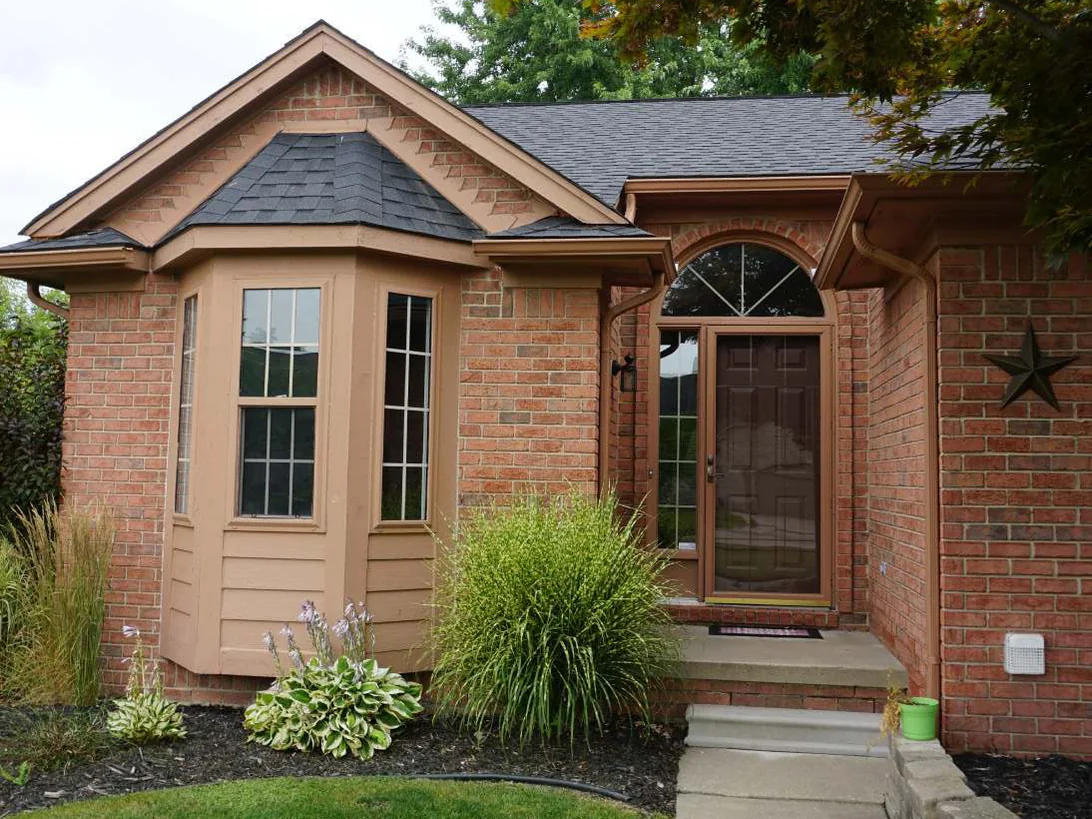 The front entrance of the house in 2019, a brick bay window with tan trim beside an arched front door, grasses and hostas in the bed and a stone-edged walk to the step
