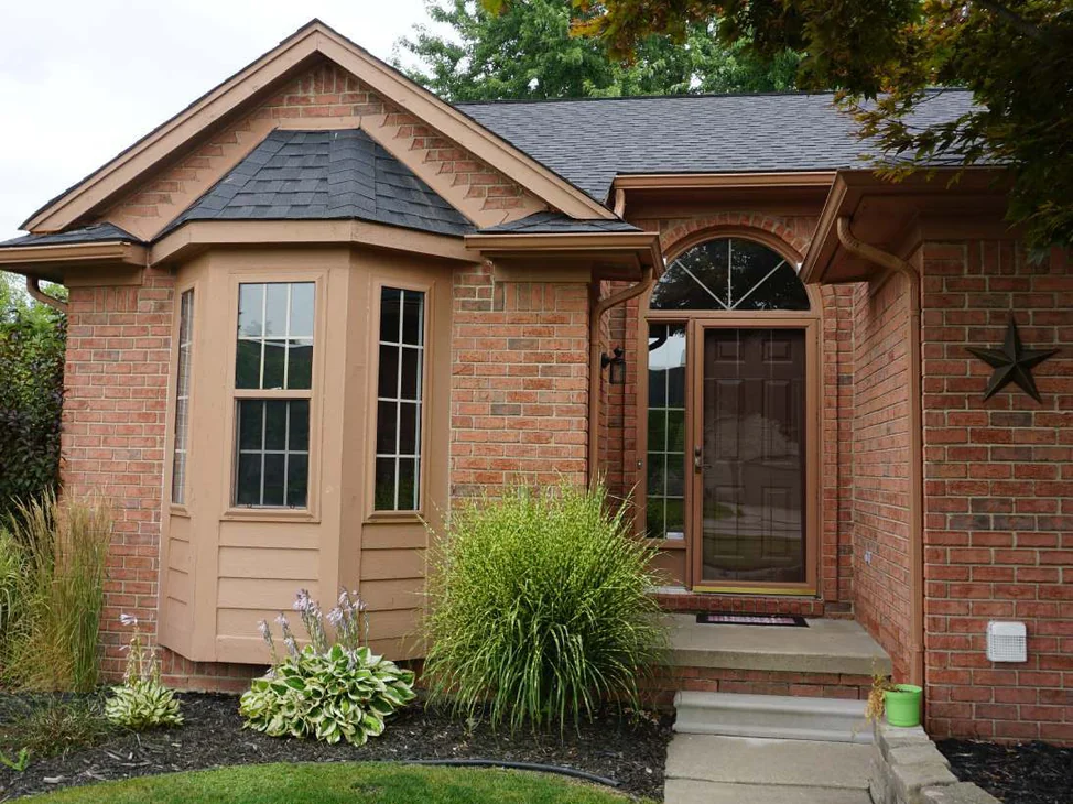 The front entrance of the house in 2019, a brick bay window with tan trim beside an arched front door, grasses and hostas in the bed