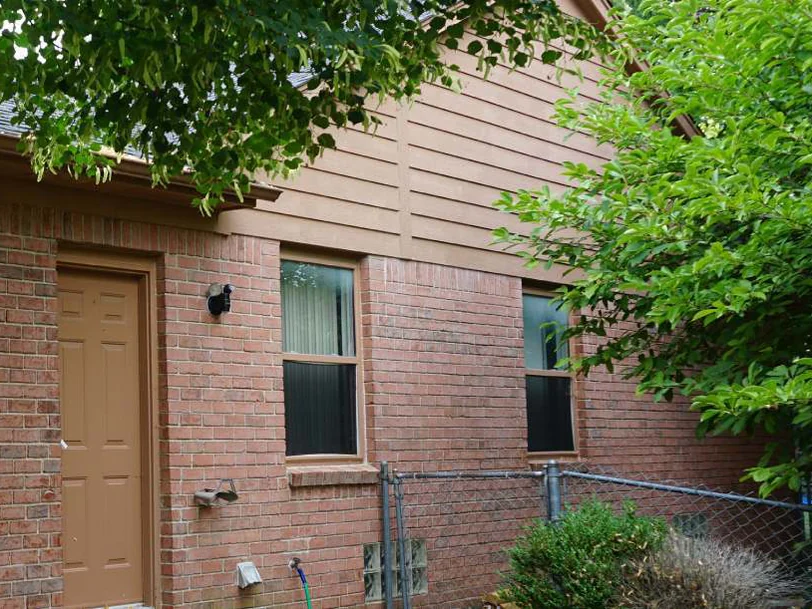 The same side wall in 2019, a tan door and two windows in even brick under the siding gable, a tree and a chain-link fence beside it