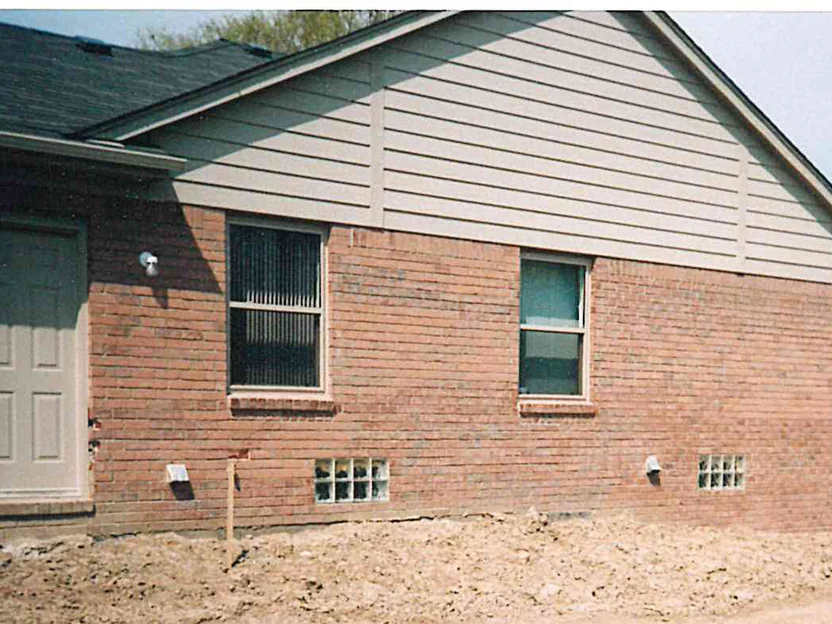 The side of the house in 1998, a siding gable over a brick wall with a door and two windows, raw earth banked at the foundation