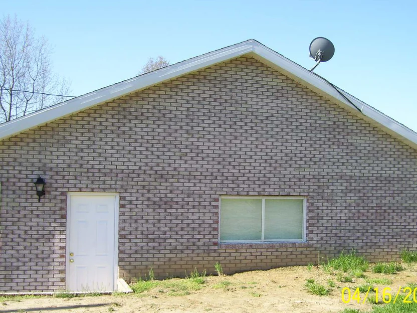 The gable end of the house in 2008, brick reading dark red and purple in patches under a satellite dish, with bare graded dirt along the base and a burned-in camera date in the corner