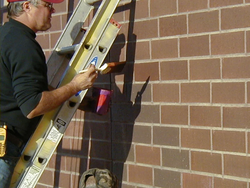 An applicator on a ladder in 2002 working stain into one brick with a hand brush, a small pot hooked to the rung beside him
