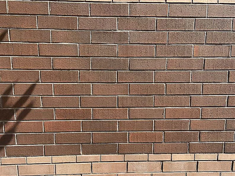The same brick at close range in 2019, red and brown units varying course by course above the cream base course, a downpipe shadow across the left of the frame