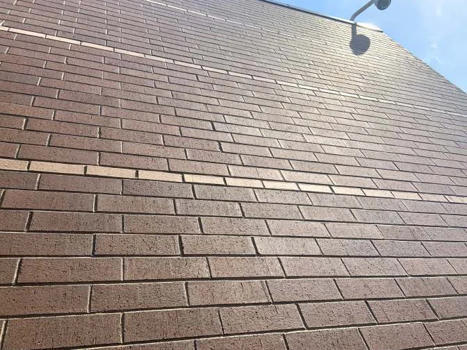 Looking up the wall in 2019 to a gooseneck light under the roofline, the brick holding one range from the ground courses to the top