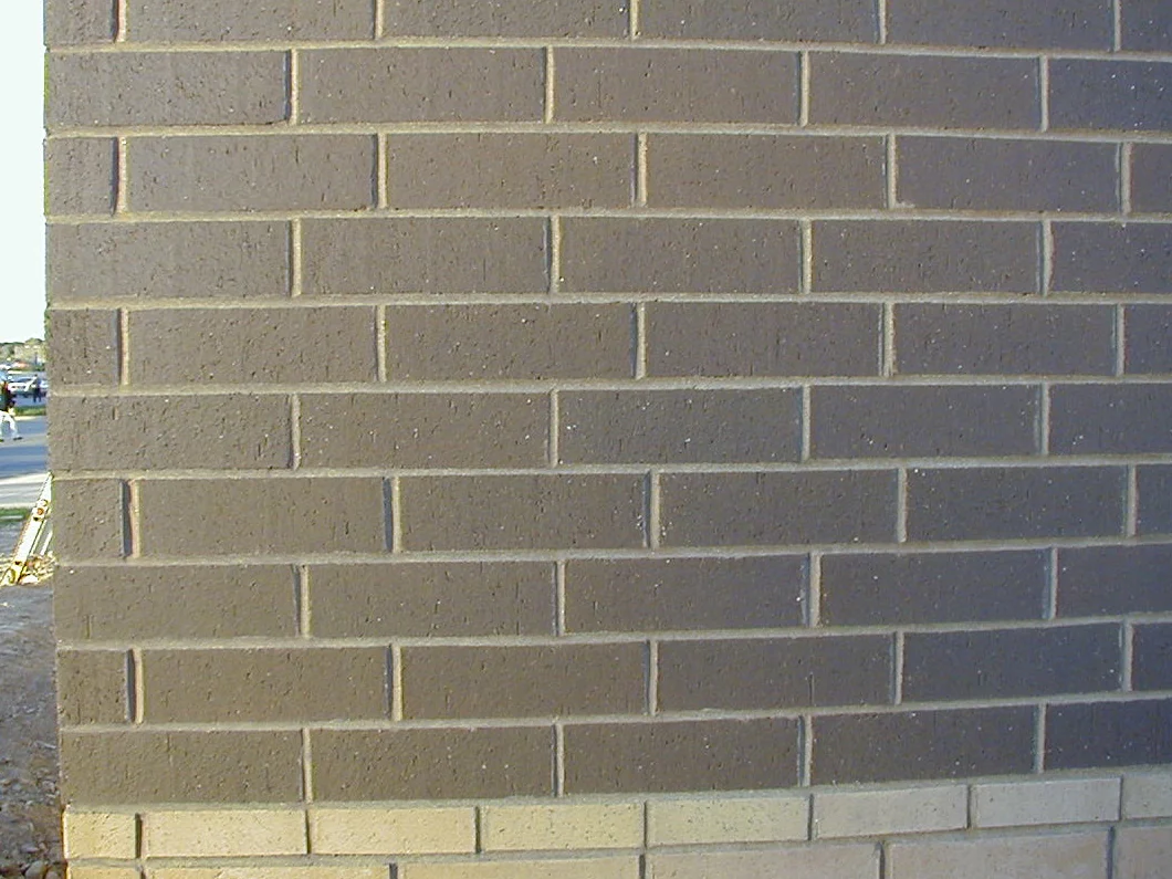 The new brick at close range in 2002, grey brown units running dark across every course above a cream base course, with bare graded dirt and a parking lot beyond the corner