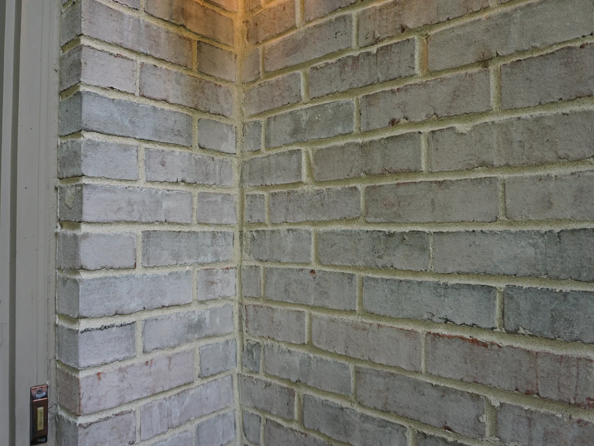 Close view of an outside brick corner beside a door frame, the units in even buff and grey tones with faint red flecks and pale mortar joints