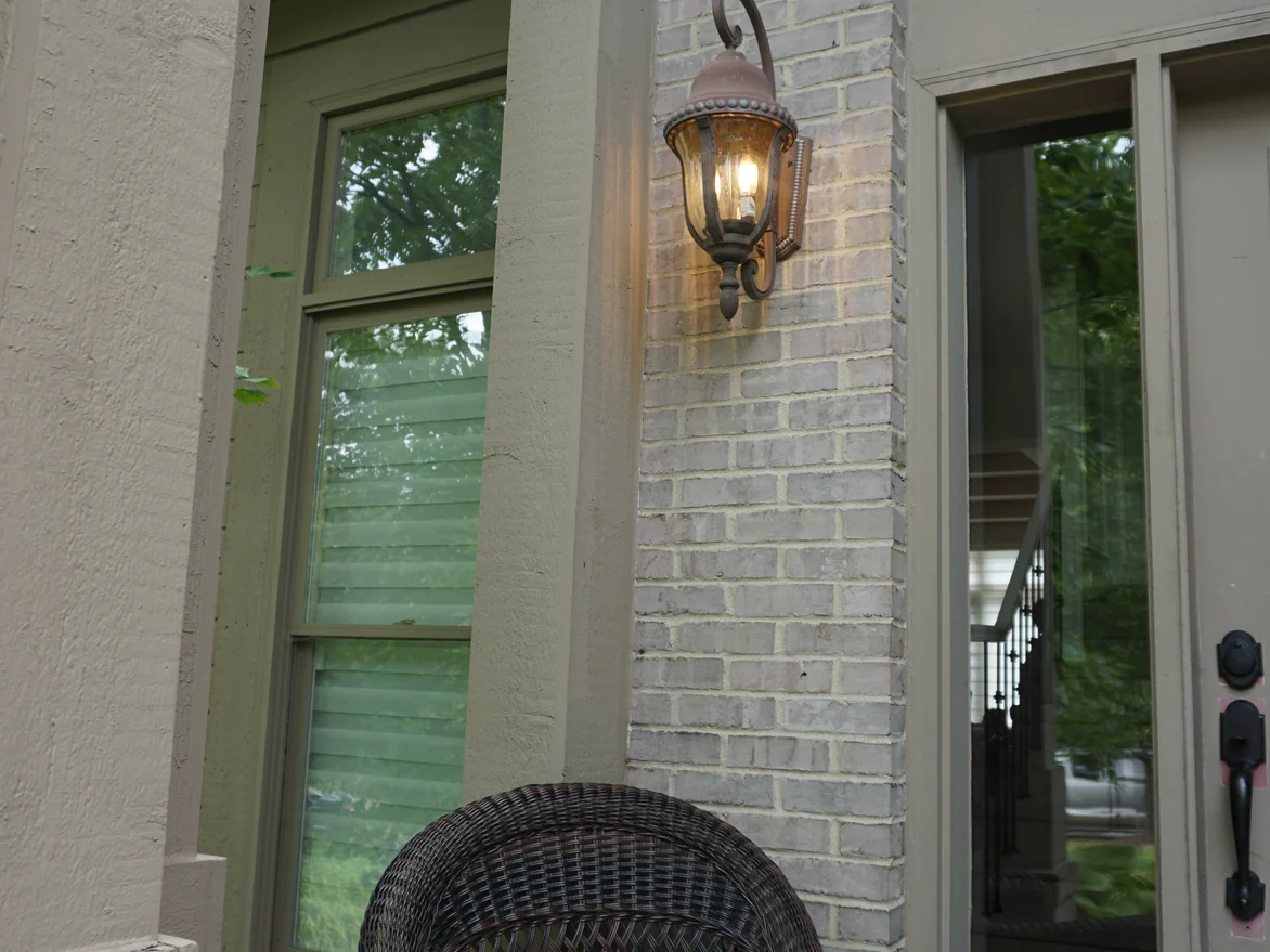 The front entry in 2019, a lit lantern on the buff brick pier beside the glazed door with a wicker chair on the porch