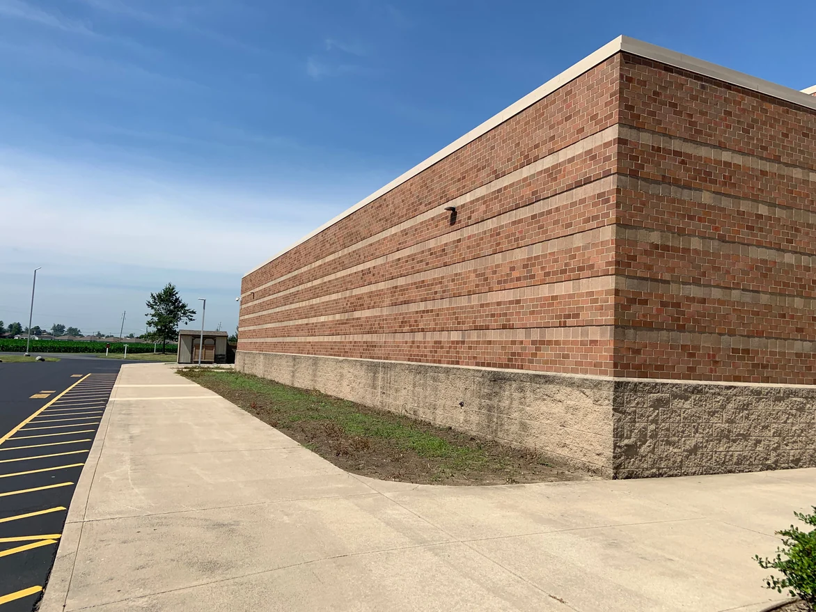 A school wall running the length of the frame to an outside corner in 2019, its horizontal accent courses even along the whole run, farm fields behind