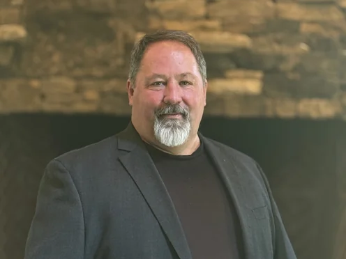 Jason McCoy in a grey blazer in front of a stone fireplace