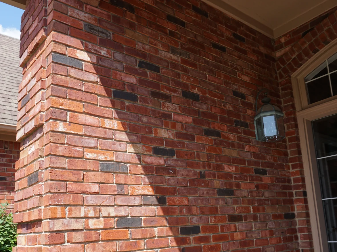 The brick pier and side wall inside the covered entry in 2019, a lantern on the wall and dark accent units running through the courses