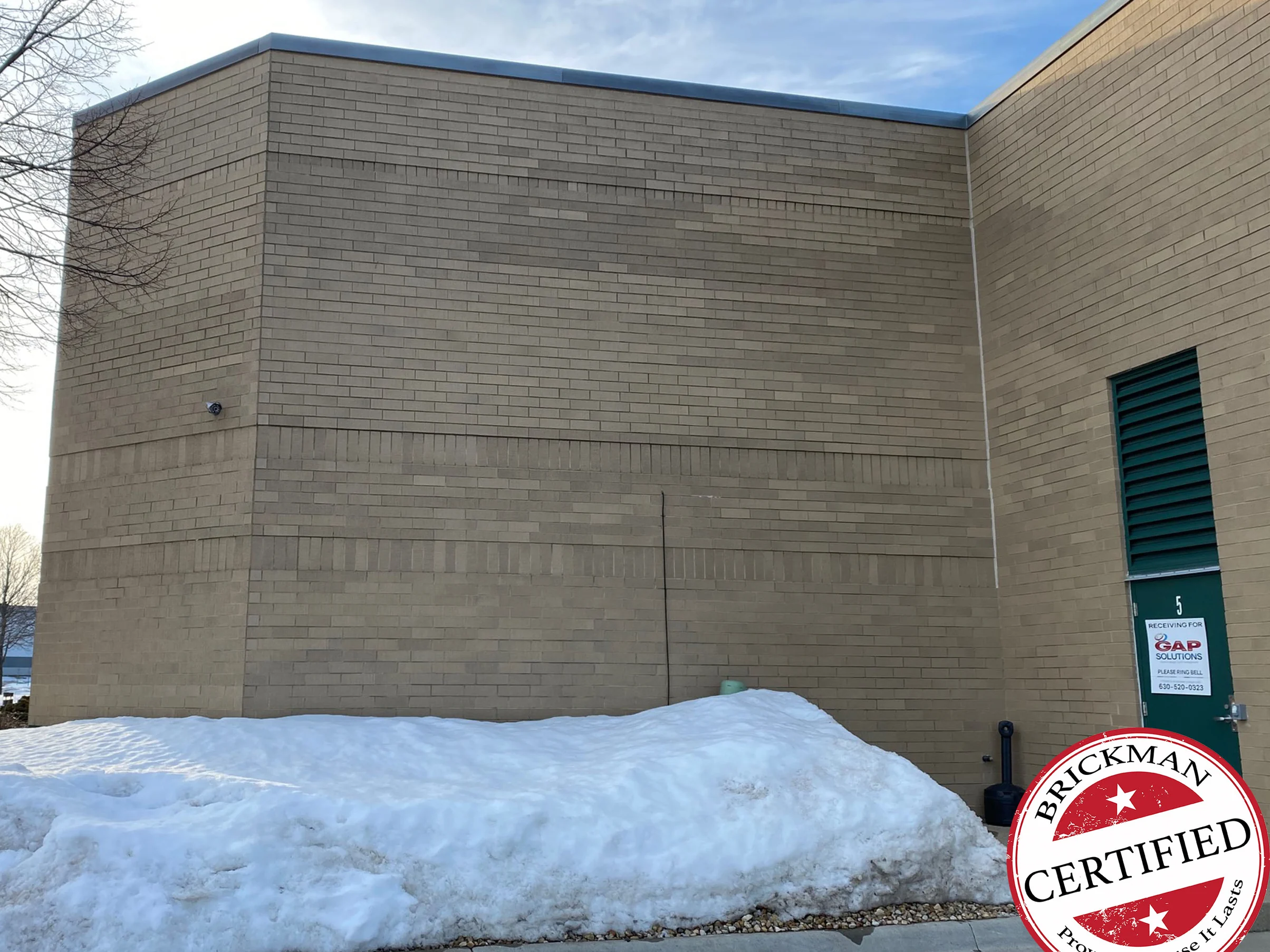 The corner of the building in 2021, buff brick with two bands of soldier courses running around it to a green door with a louver above, snow banked at the base and the certification stamp in the corner