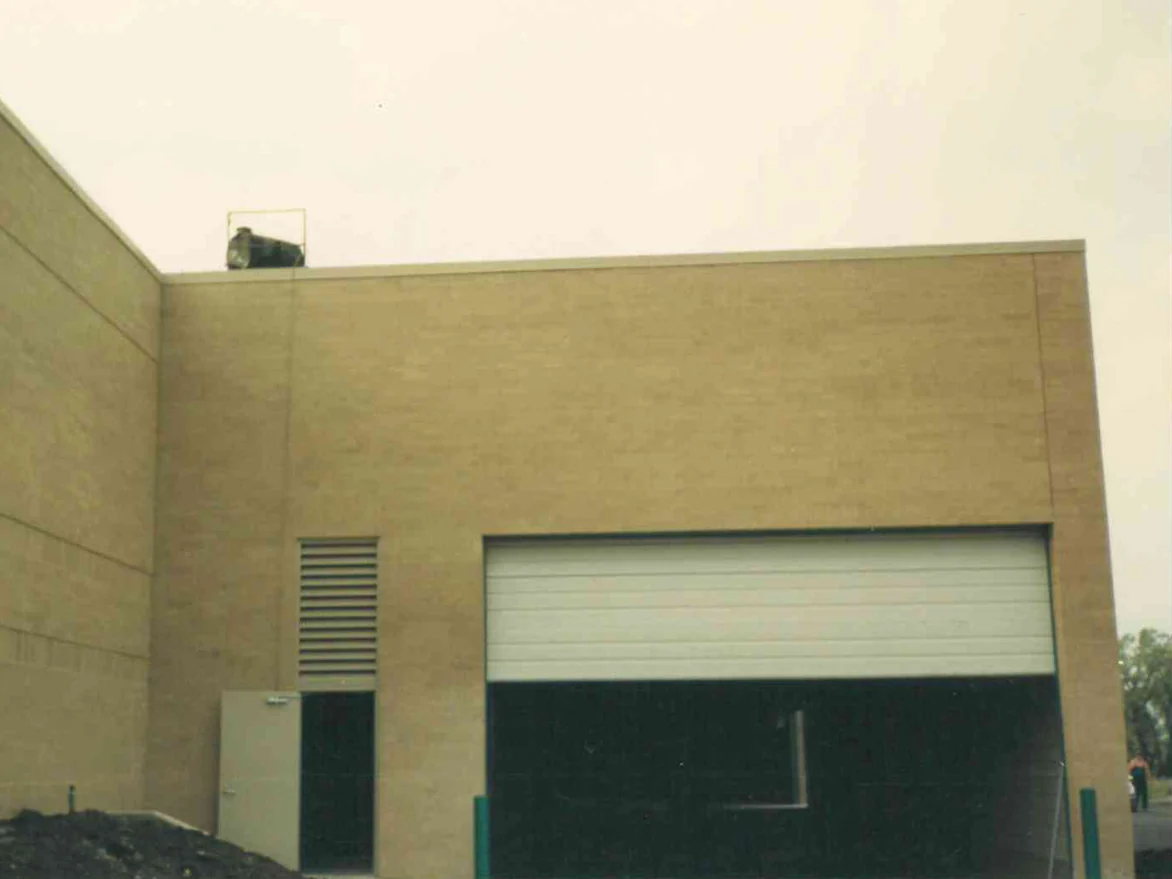 A buff brick wall in 1991 beside a taller wing, with a louver, a metal door and an open overhead loading door, darker blotches drifting across the courses and a pile of fill in front