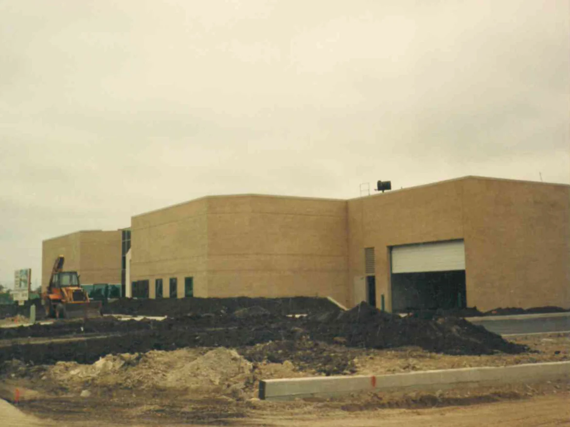 The whole building from across the graded site in 1991, buff brick blocks stepping back to an office wing with a ribbon of windows, a backhoe and heaped soil in the foreground
