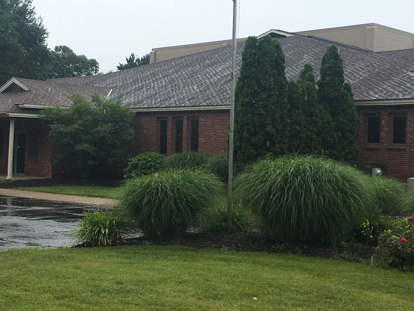 The long side elevation in 2019, red brick and tall narrow windows under the shingle roof, a flagpole in front and ornamental grasses and evergreens against the wall