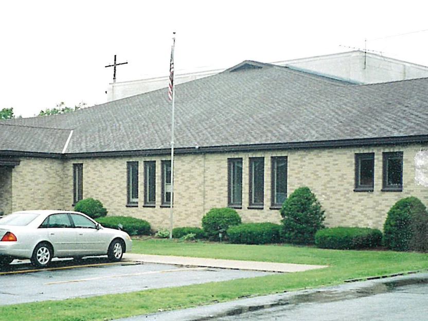The long side elevation of the church in 2001, a low buff brick wall of tall narrow windows under a wide shingle roof, with a flagpole and a cross on the roofline above