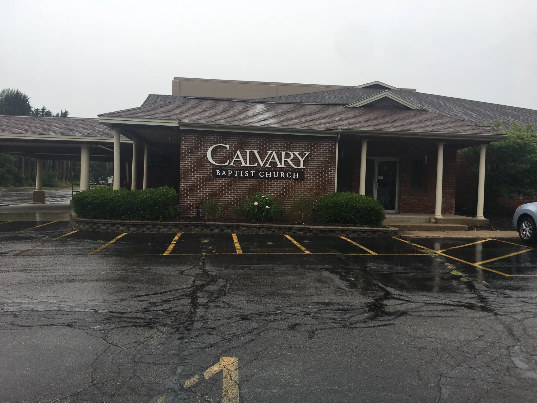 The church entrance in the rain in 2019, red brick under a drive-through canopy on round white columns, a sign reading Calvary Baptist Church on the wall and clipped hedges along the base