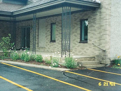 The church entrance in June 2001, buff brick with darker units scattered through it under a porch roof on black iron trellis columns, daisies in the bed and a camera date of 6 21 01 in the corner