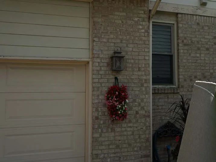The brick pier between the garage door and the window in 2019, a black lantern and a wreath of pink flowers on it, the brick one even blend