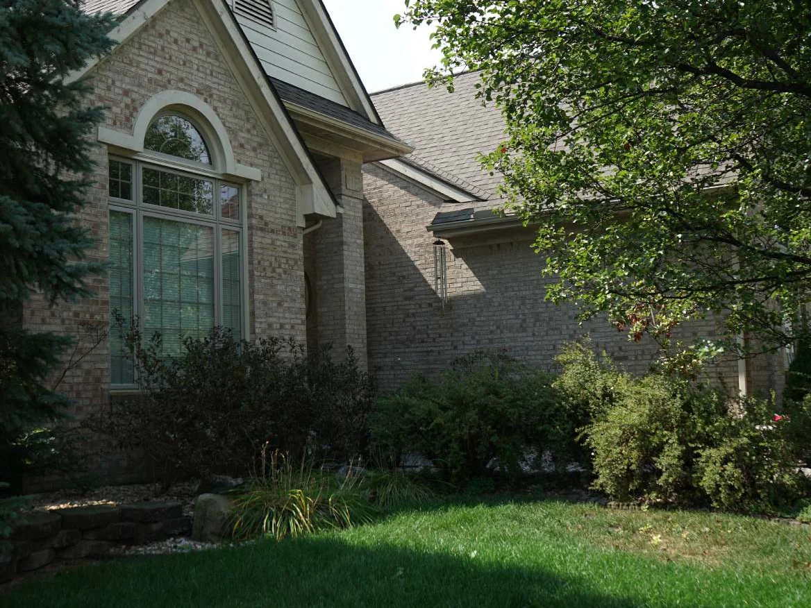 The front gable of the house in 2019, an arched window in cream and tan brick under a siding peak, a spruce on the left and a shade tree over the lawn