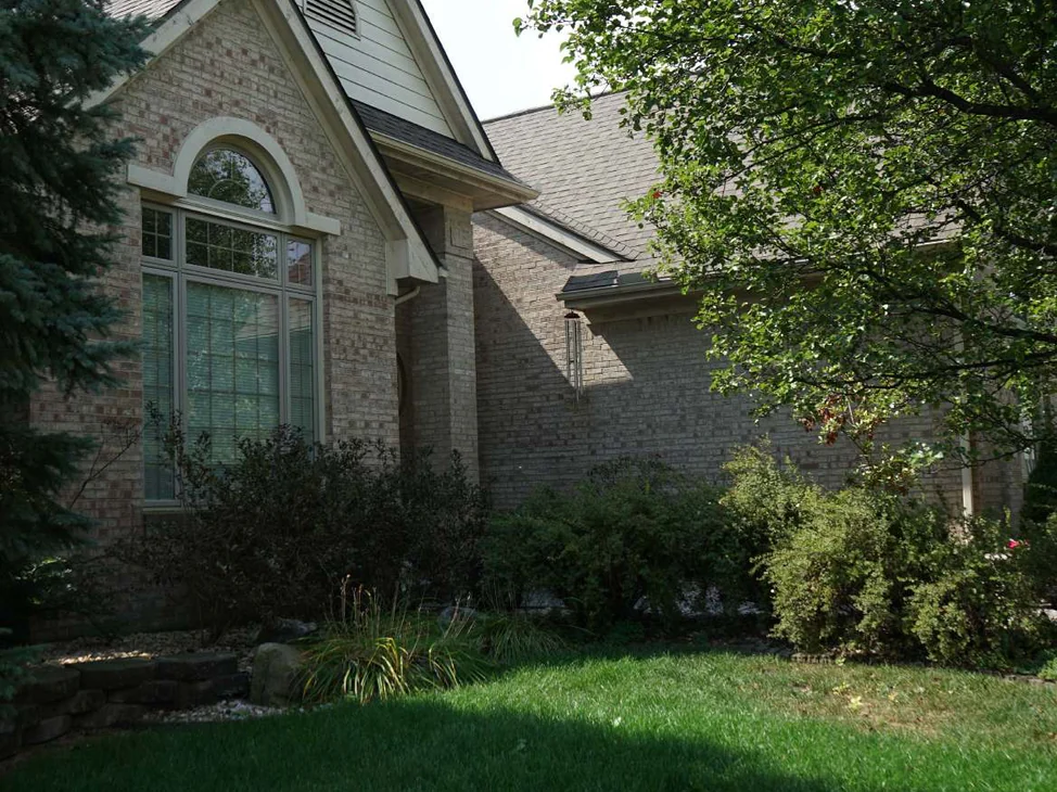 The front gable of the house in 2019, an arched window in cream and tan brick under a siding peak, a spruce on the left and a shade tree over the lawn
