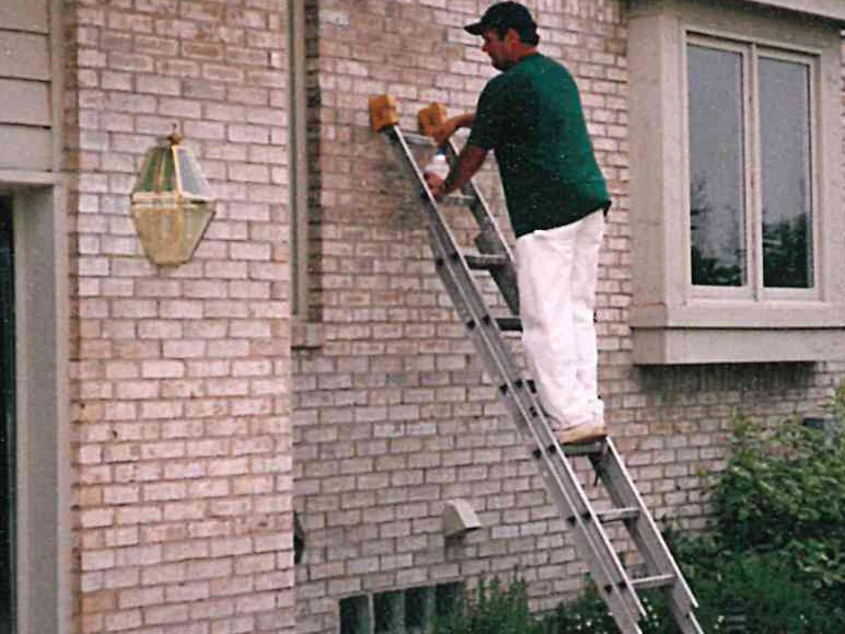 An applicator on an extension ladder in 1997 working stain into the wall beside the bay window with a hand brush, a lantern on the pier below him