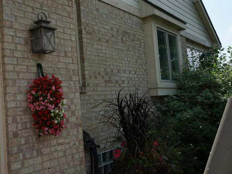 The wall beside the bay window in 2019, a black lantern and a hanging basket of pink flowers on even cream and tan brick