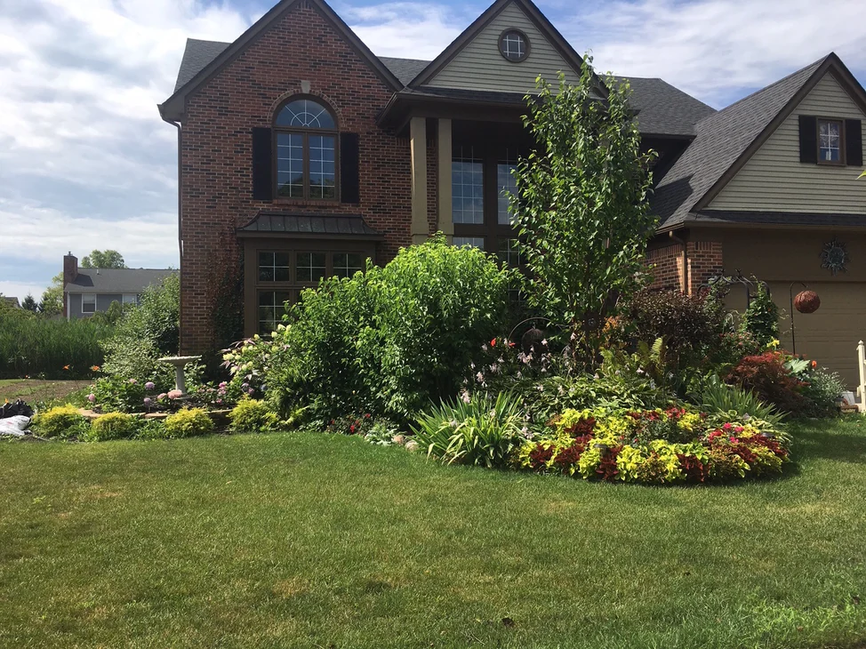 The front of the home from across the lawn in 2019, its brick one red and brown range behind deep planted beds and a young tree.