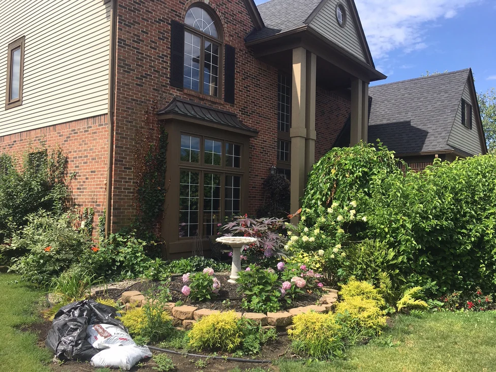 The front corner of the home in 2019, a birdbath and planted beds standing against the brick below the bay window