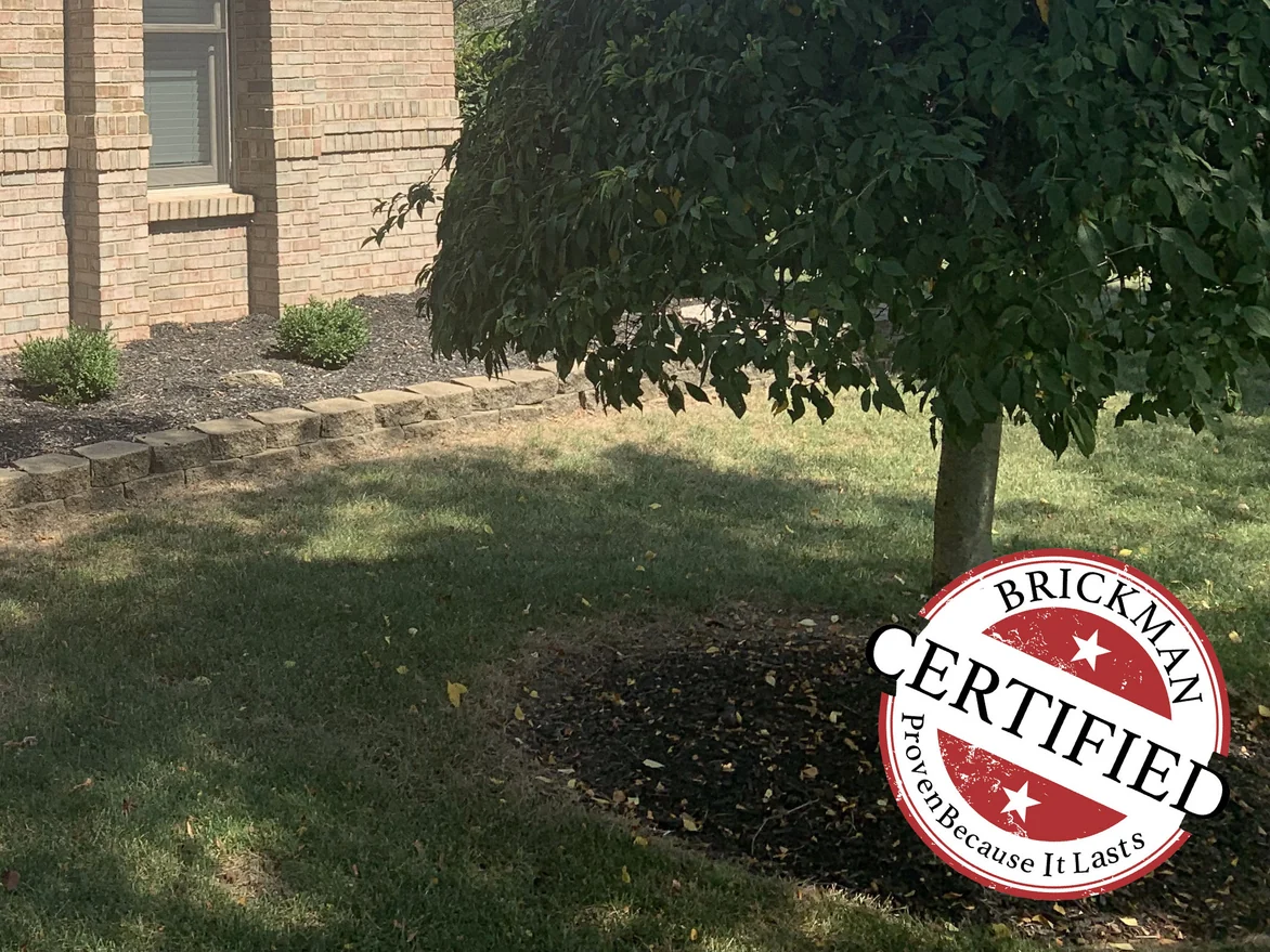 The end wall of the front room in 2019, its window and brick standing behind a clipped hedge, with the Brickman certification stamp over the lawn