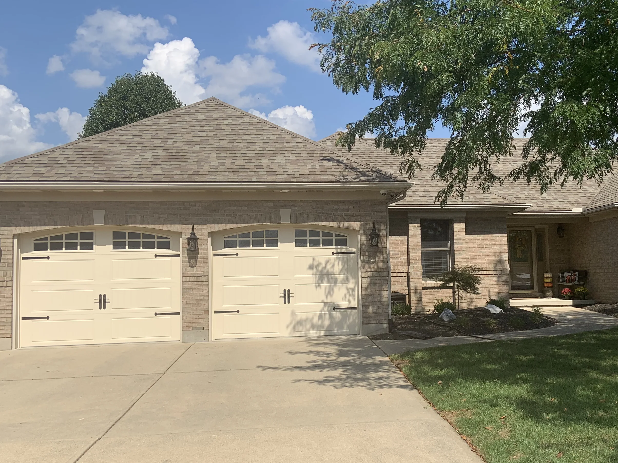 The front of the house from the driveway in 2019, the garage bays, the entry and the hipped roof over one even cream and tan brick range