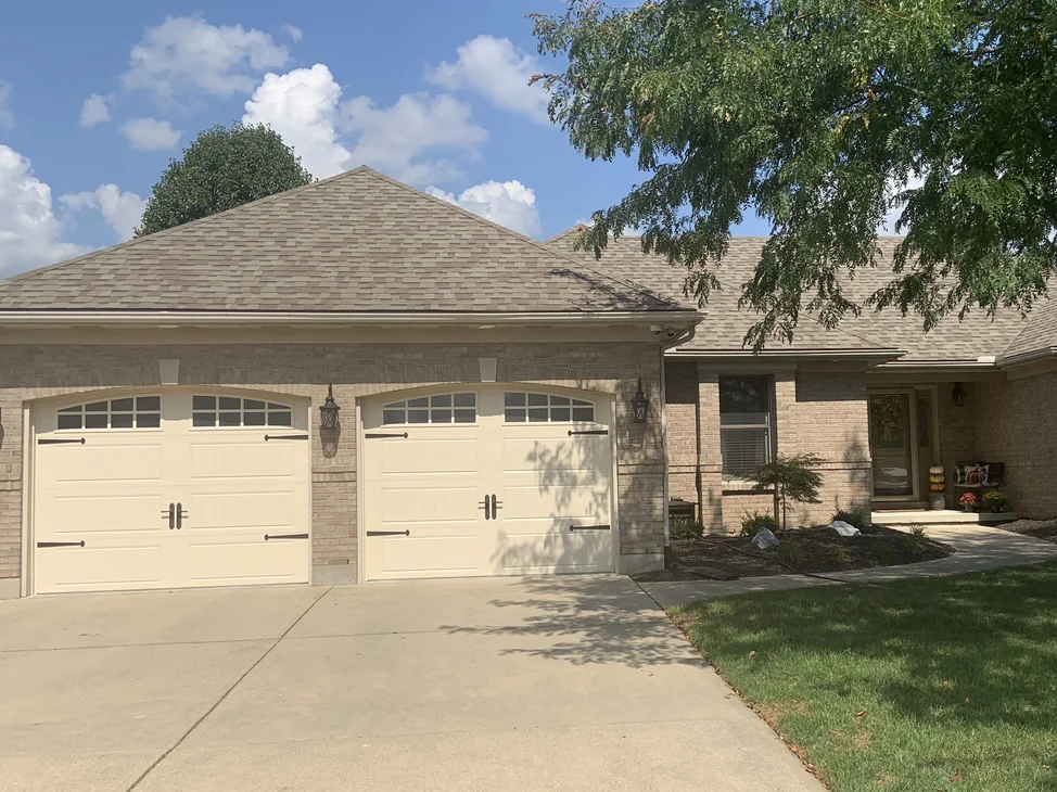 The front of the house from the driveway in 2019, the garage bays, the entry and the hipped roof over one even cream and tan brick range.