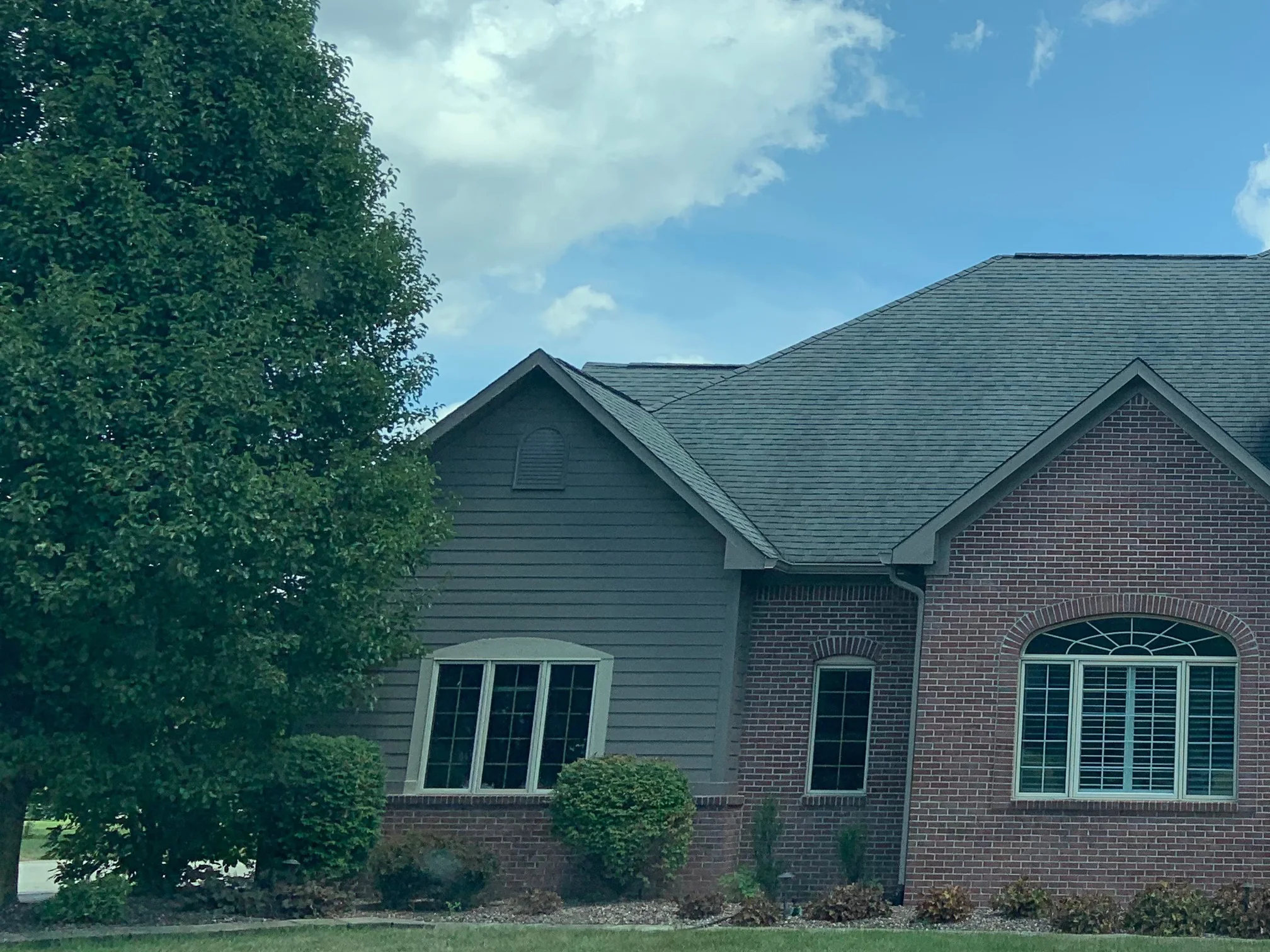 The house from the street in 2019, its brick gable and sided wing under one roofline behind a mature front tree
